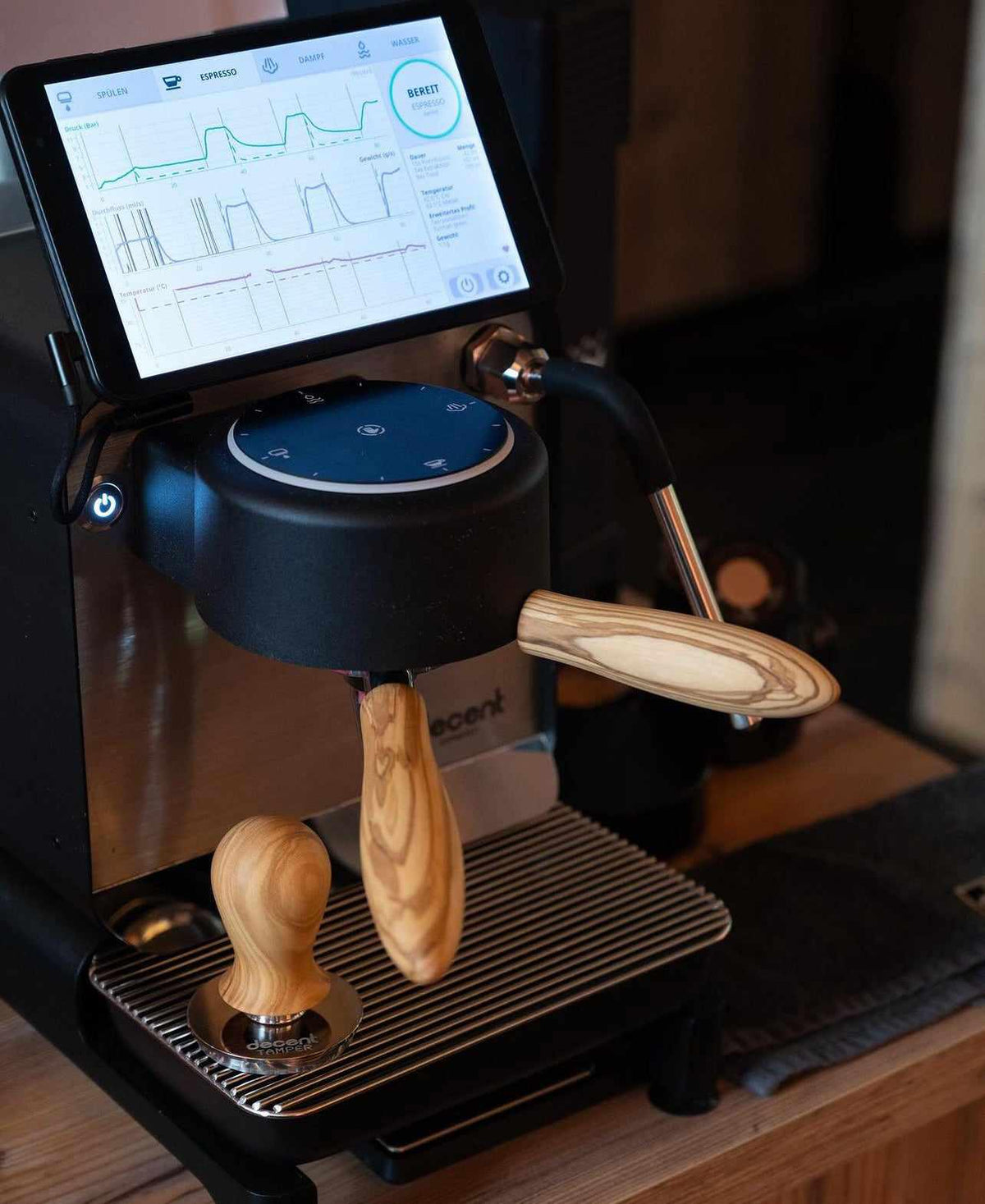 Espressomaskin med digital skärm som visar bryggningsstatistik och en tamper med trähandtag på avläggningsplattans yta.