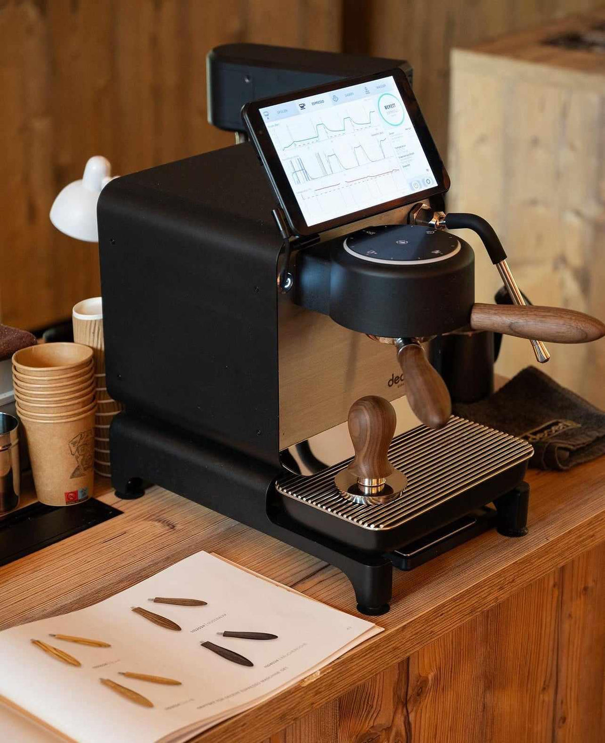 Espressomaskin med digital skärm och trähandtag placerad på träbord vid papperskoppar och prov på kaffeverktyg.